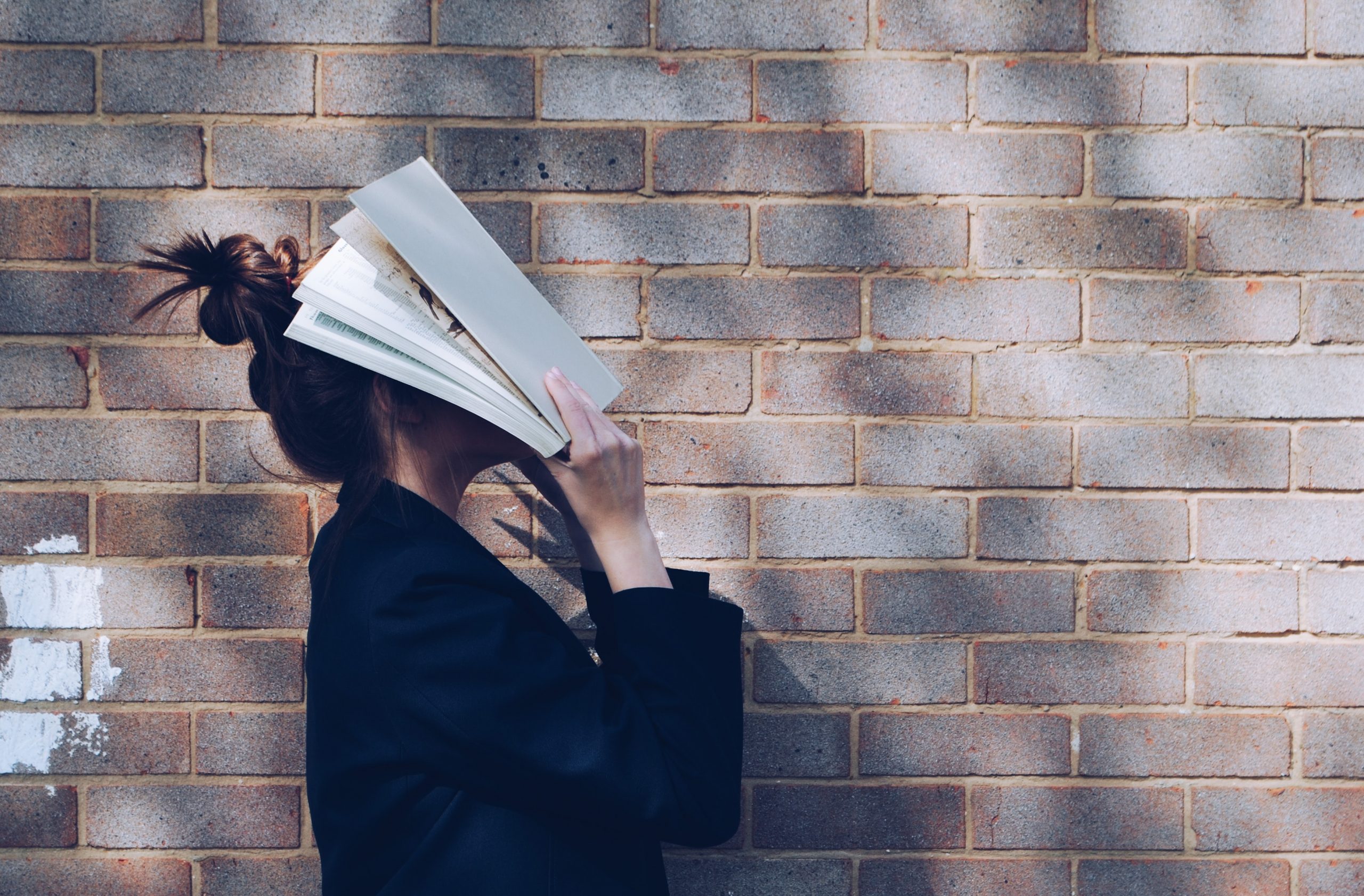 Jeune femme tenant son visage dans un livre ouvert, devant un mur de briques